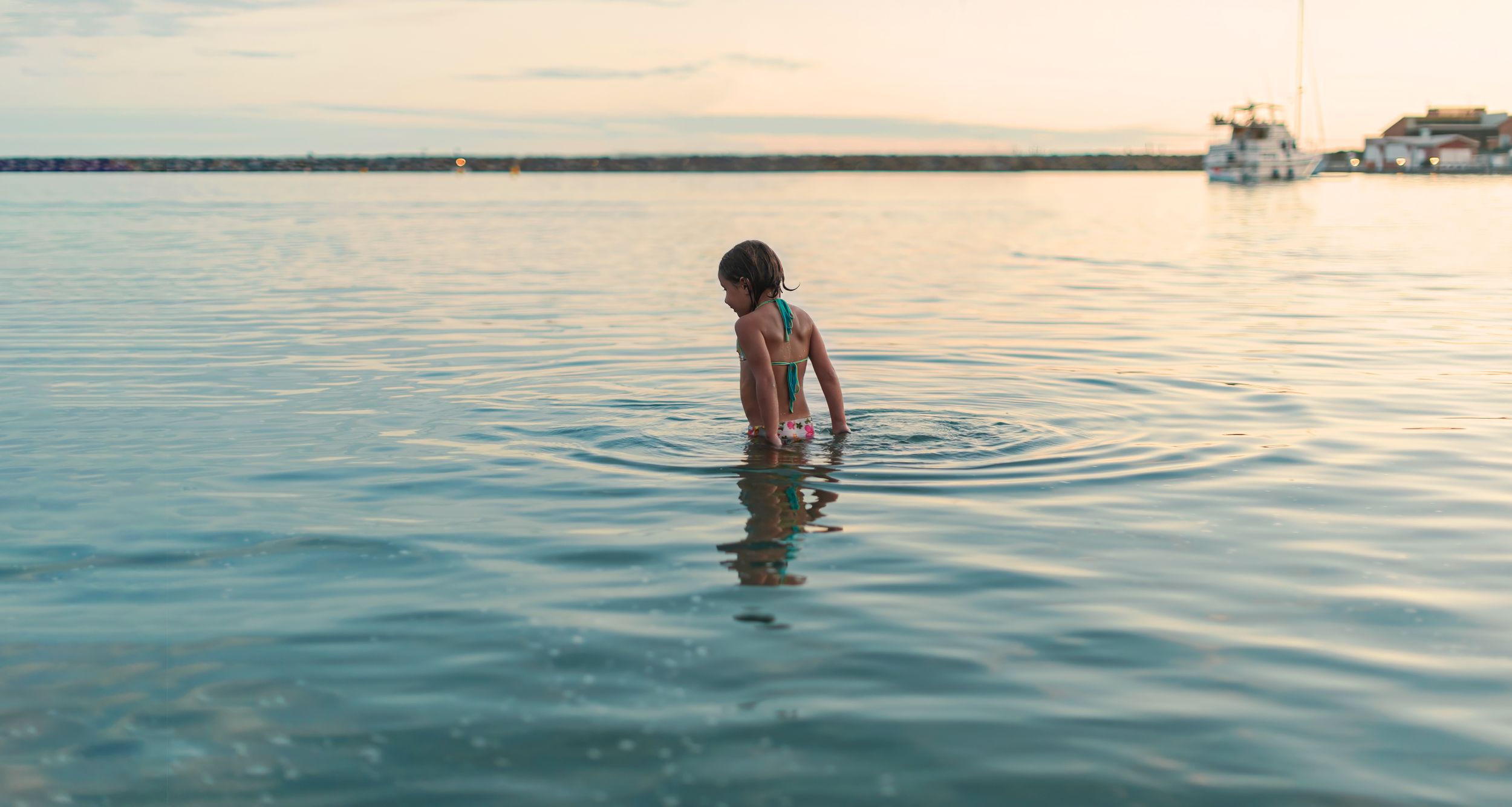 Young child wading in calm lake water at sunset with a boat visible in the background.