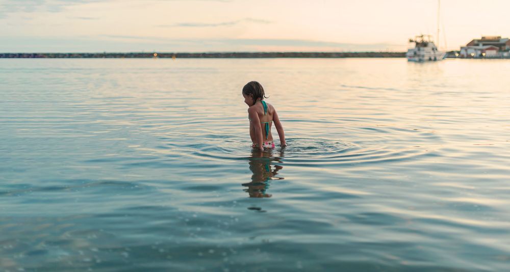 Young child wading in calm lake water at sunset with a boat visible in the background.