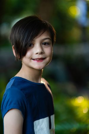Young boy with dark hair smiling softly in blue shirt against green bokeh background.