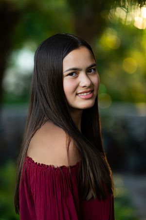 Teen girl with long dark hair smiling over shoulder in red off-shoulder top outdoors.