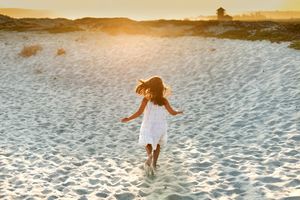 Young girl in white dress running across sandy beach toward golden sunset.