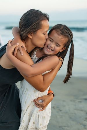 Mother kissing young daughter on the cheek while hugging on a beach at dusk.