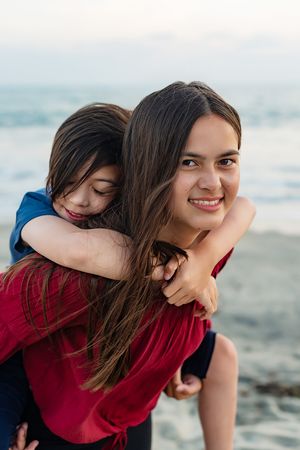 Smiling young woman giving a child a piggyback ride on a sandy beach.