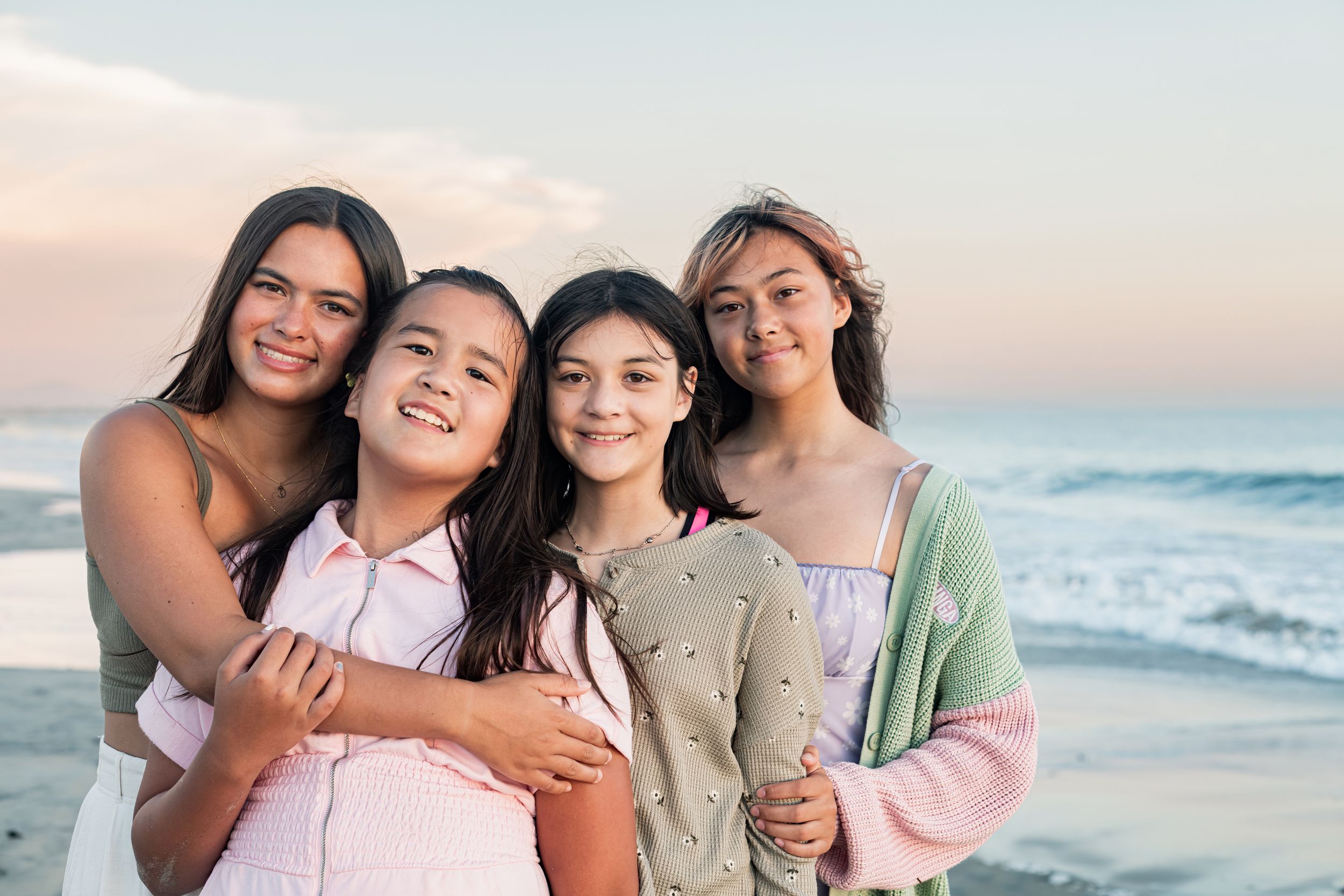 Four smiling young girls pose together on a beach during a pastel sunset, hugging warmly.