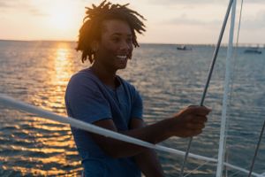 A smiling young man with dreadlocks holding a sailboat rope at sunset over a calm ocean.