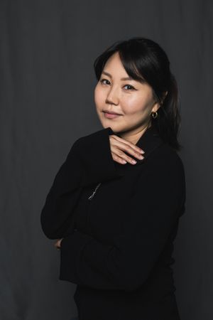 Asian woman in black turtleneck posing with arms crossed against a grey studio background.