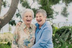 Two elderly women laughing and embracing outdoors under a tree with ocean view in background.