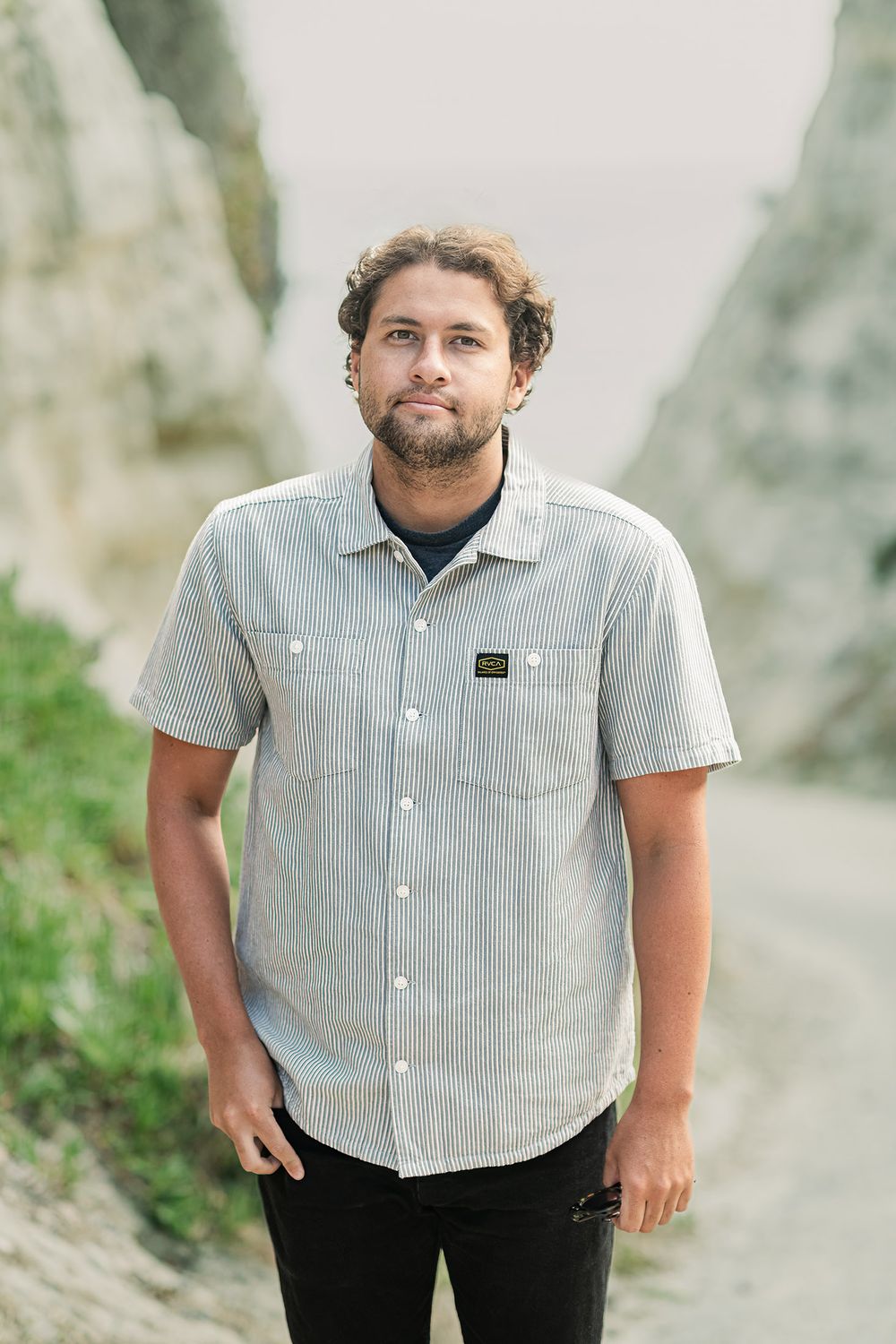 Young man with beard wearing striped shirt standing between rocky coastal cliffs.