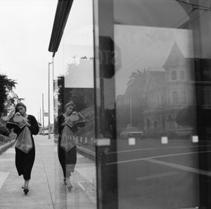 Black and white photo of a woman walking past a reflective storefront, mirroring a Victorian house and street scene.