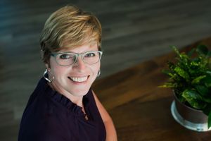 Smiling woman with short blonde hair and glasses poses at a wooden table with a green plant nearby.