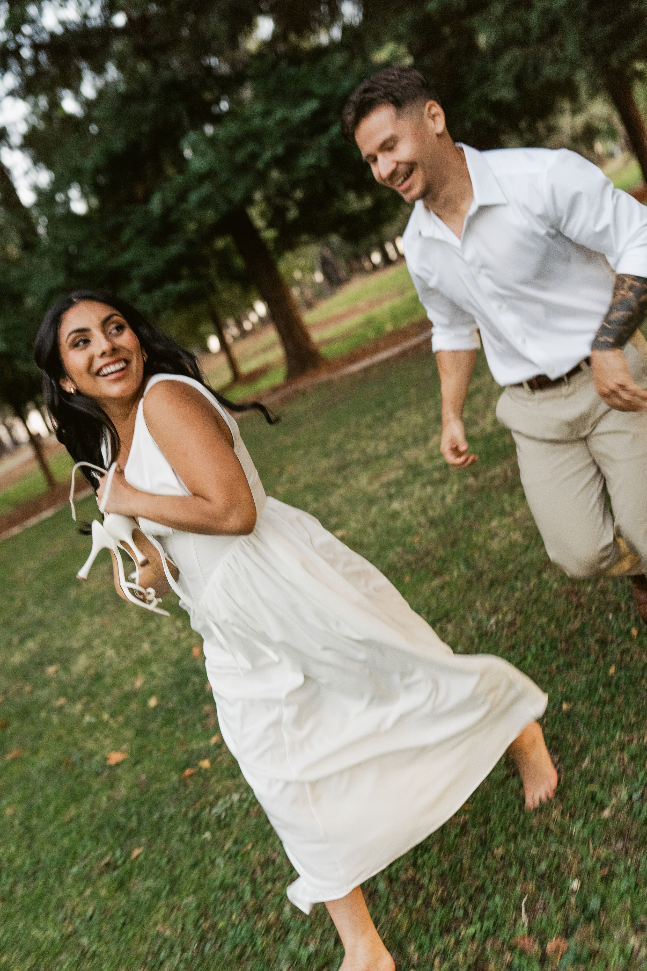 A couple in white and beige outfits playfully dance and twirl together on a grassy lawn in a romantic photo series.