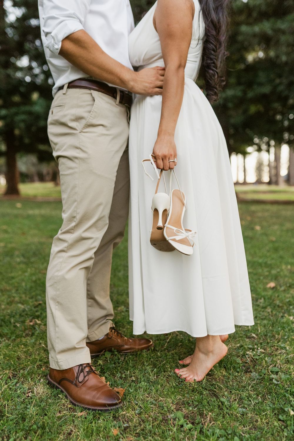 Close-up shot of feet showing brown leather dress shoes and white heels on grass in an outdoor setting.