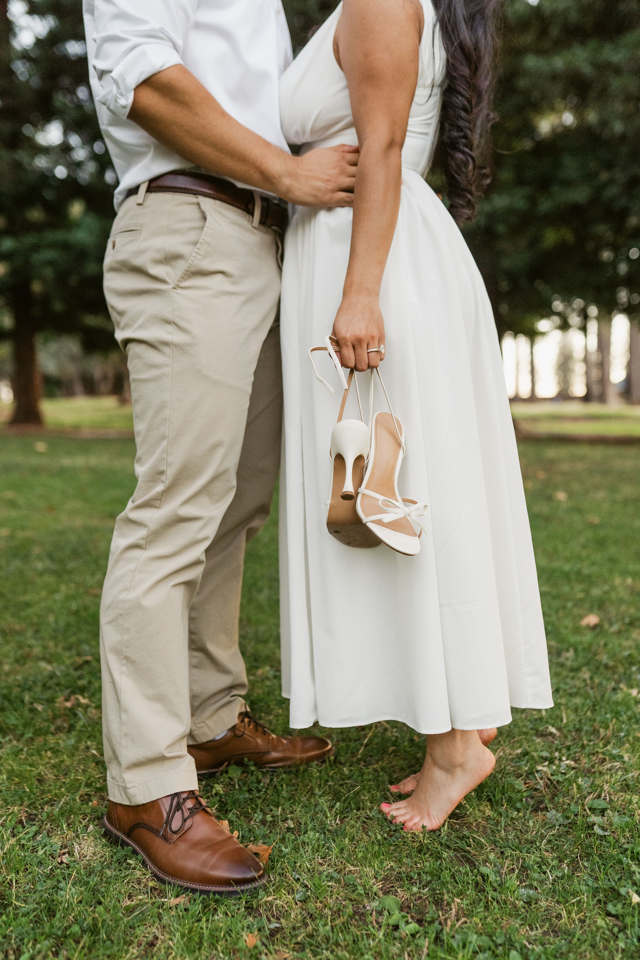 Close-up shot of feet showing brown leather dress shoes and white heels on grass in an outdoor setting.