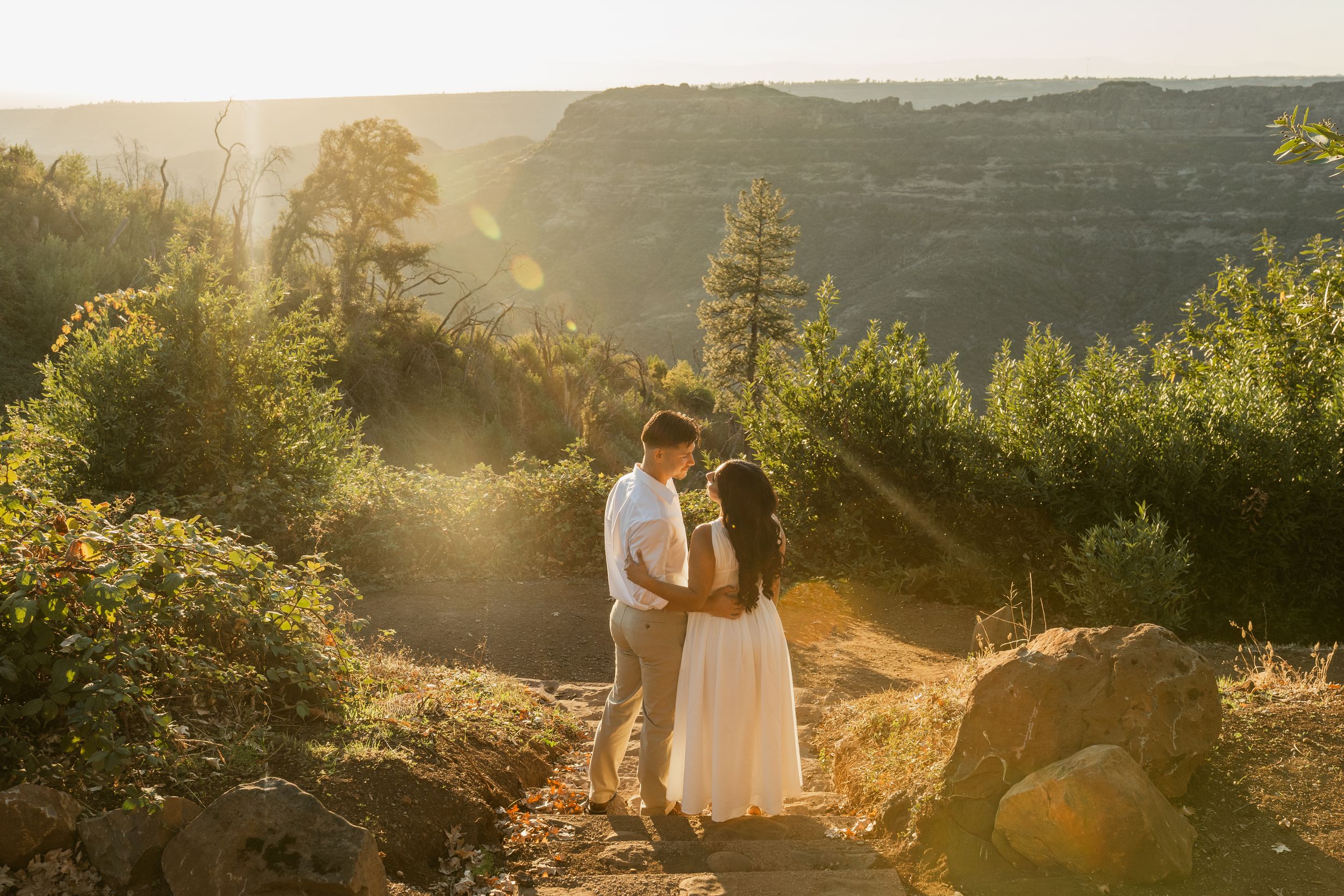 Couple embraces during golden hour photoshoot in scenic mountain landscape with dramatic sunlight and trees.