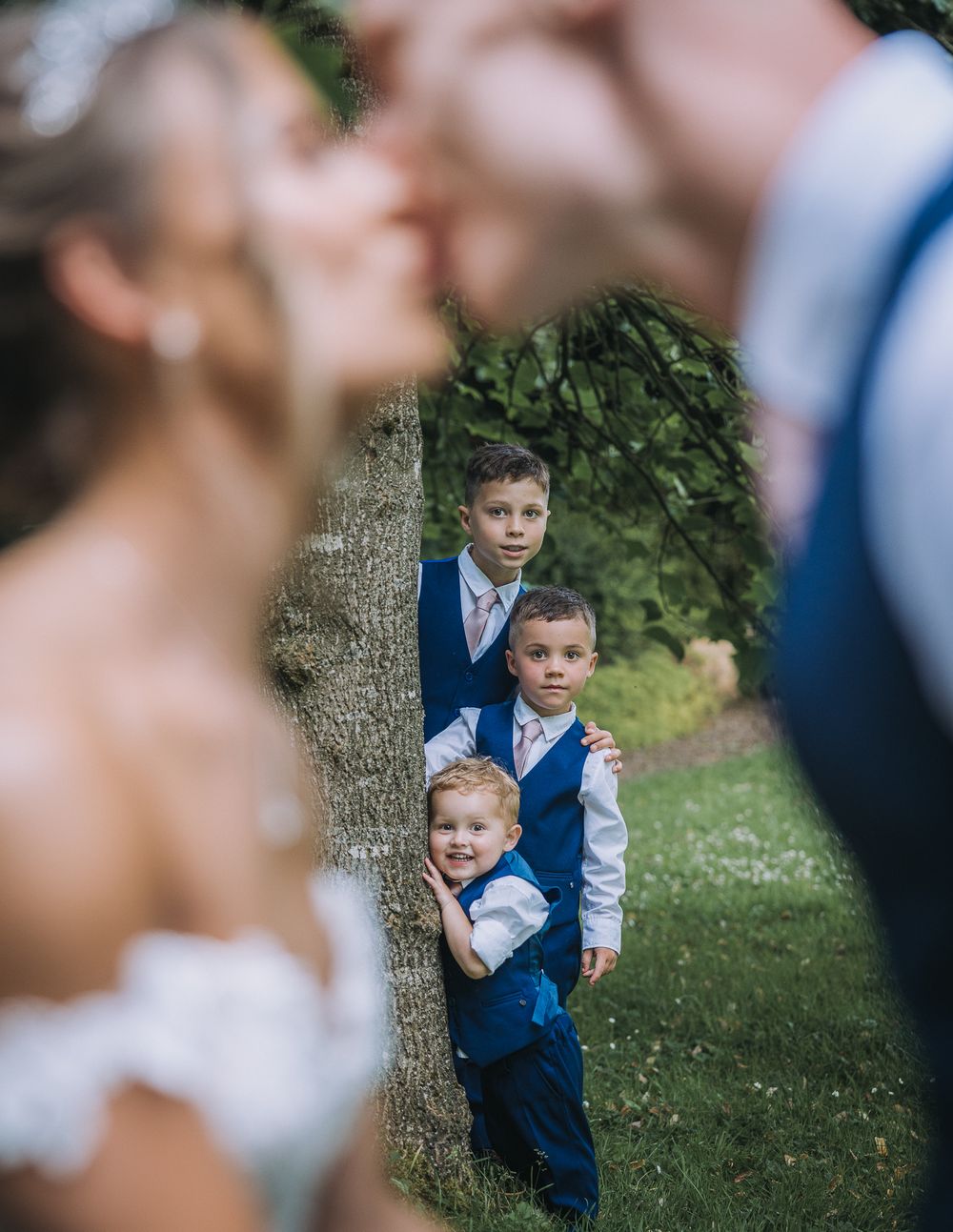 Young attendees in matching blue vests peek out from behind a tree while watching a couple in the foreground. The Mill Barns