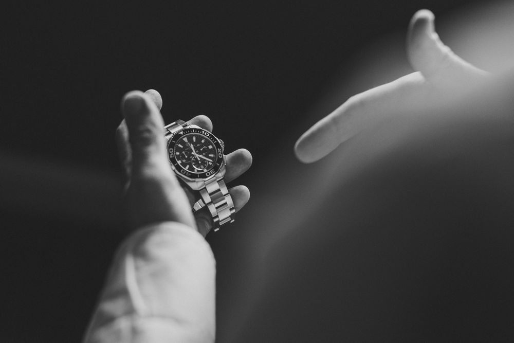 A silver wristwatch suspended in midair between two outstretched hands in a black and white photograph. Groom Prep