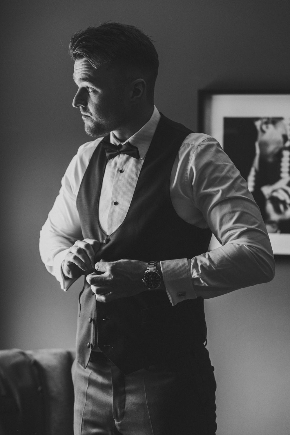 A black and white photograph of a person adjusting their formal vest while wearing a bow tie and dress shirt. Groom Prep