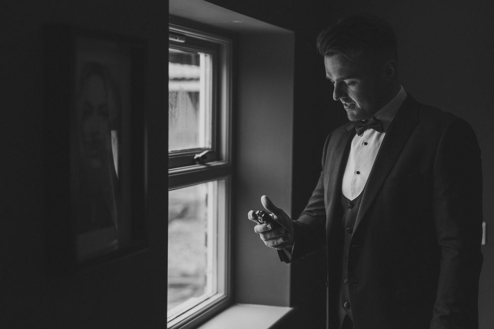 A person in a tuxedo stands by a window in dramatic black and white lighting while looking at their phone. Groom Prep