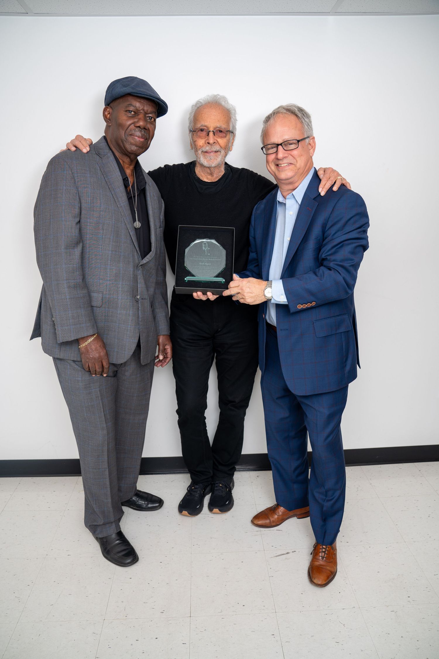 Three men in casual and formal attire pose together against a white wall for a photo shoot.