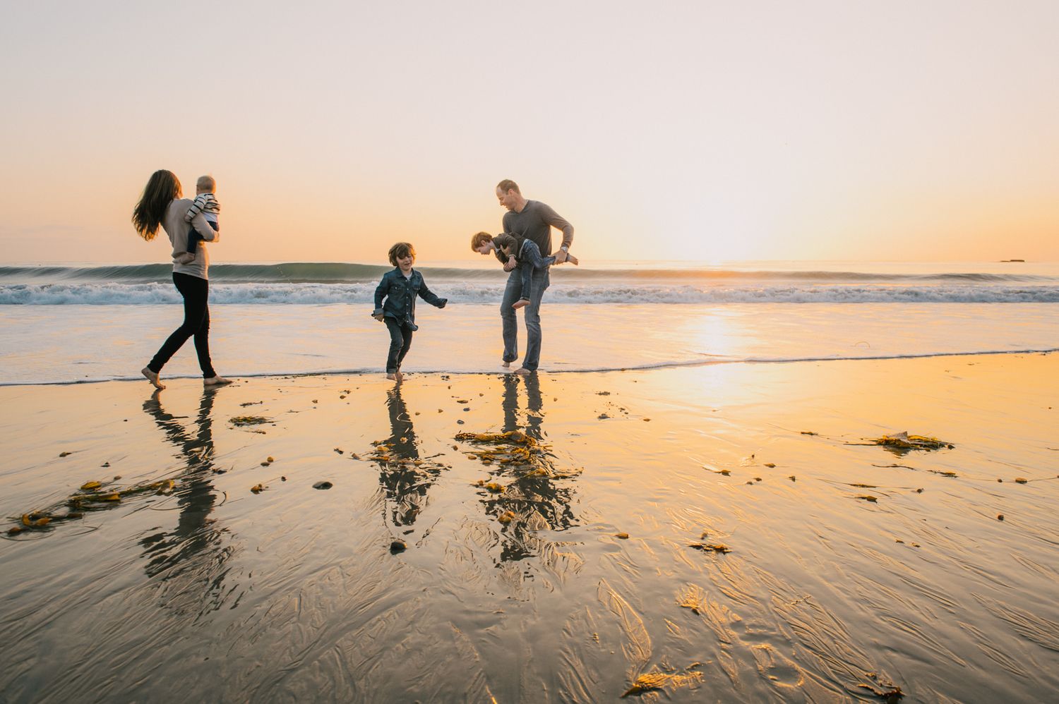 Happy family of four walks along a reflective beach shoreline during a golden sunset together.
