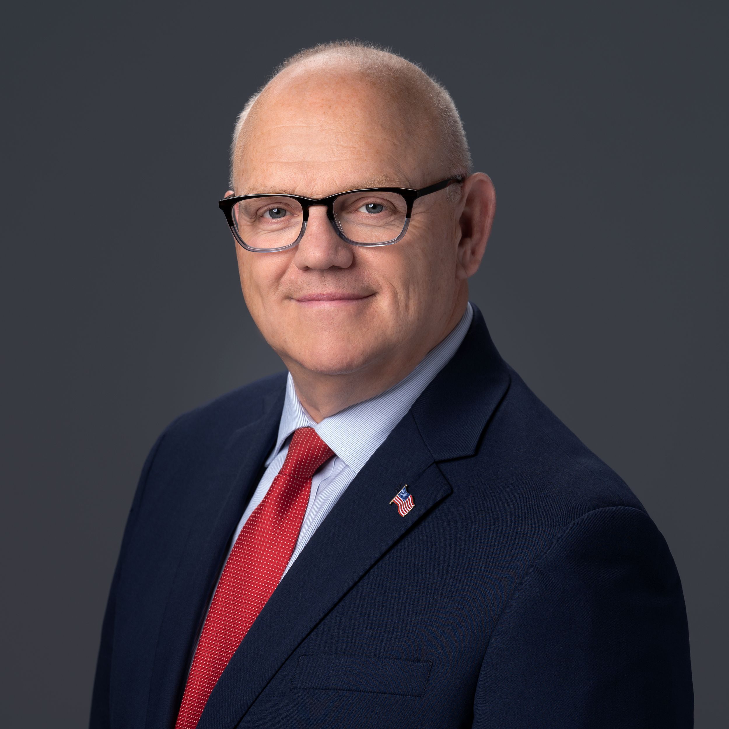 Professional headshot DA Tim Cruz in a, navy suit and red tie against dark background.