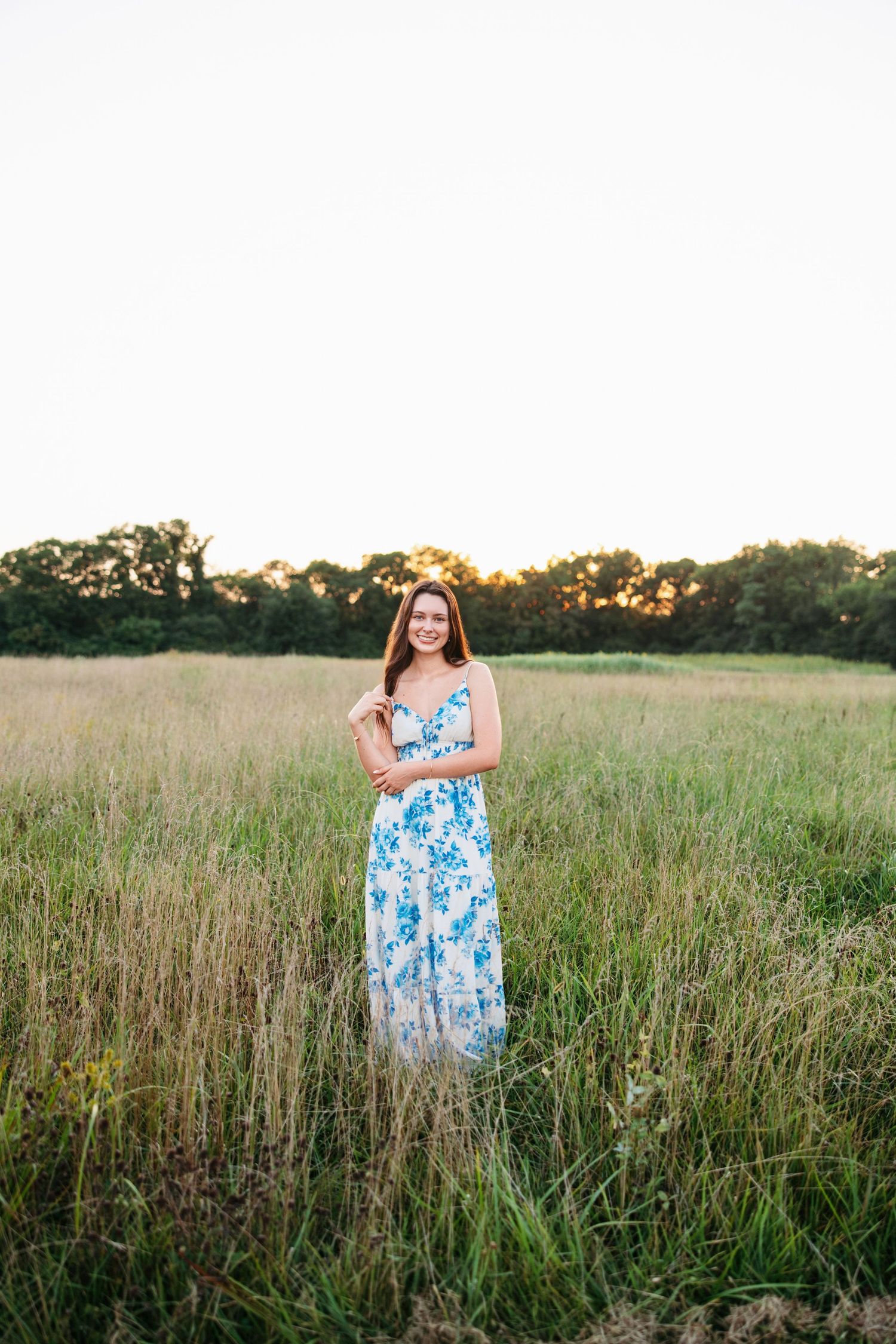 A woman in a blue and white floral dress poses gracefully in a golden field during sunset.