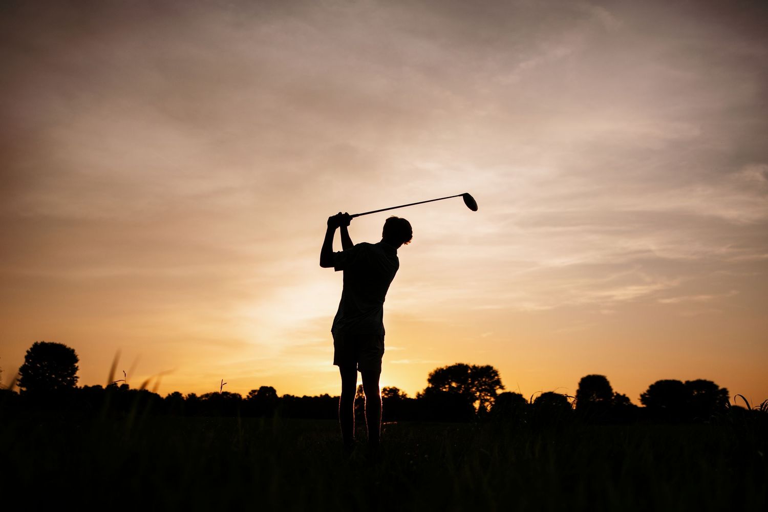 Silhouettes of golfers practicing swings against a dramatic sunset sky with trees in the background.