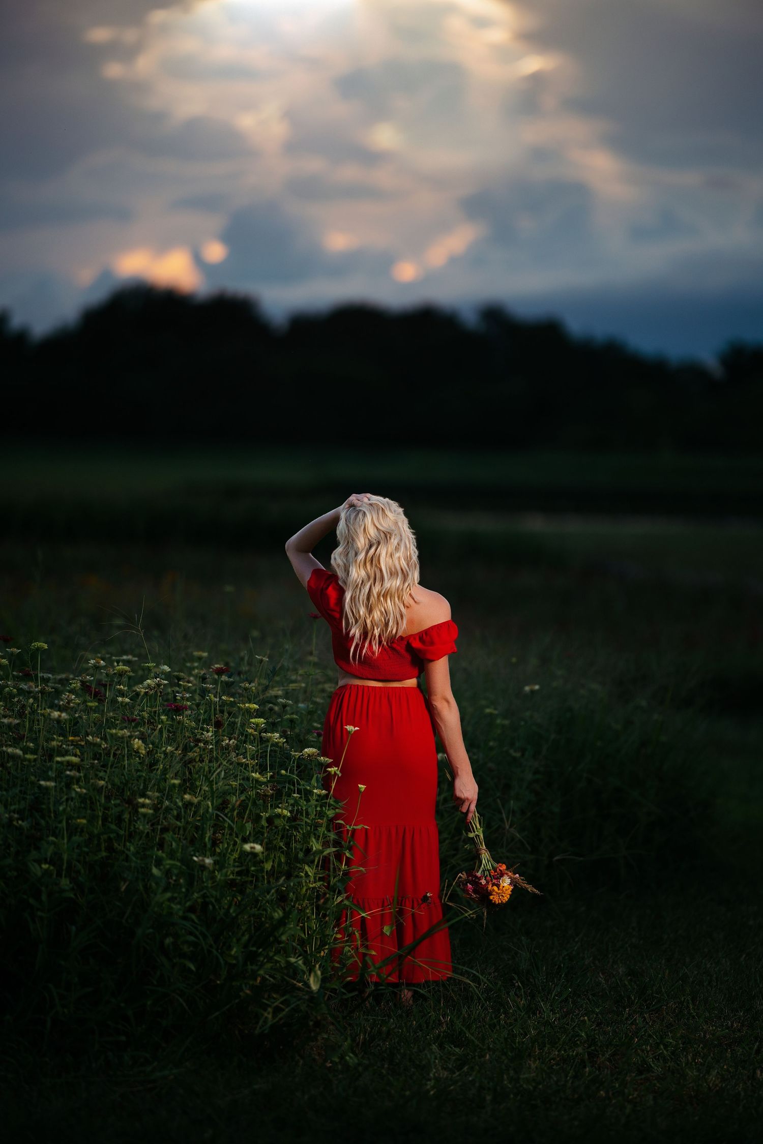 A person in a flowing red dress stands in a dusky wildflower meadow holding flowers at sunset.