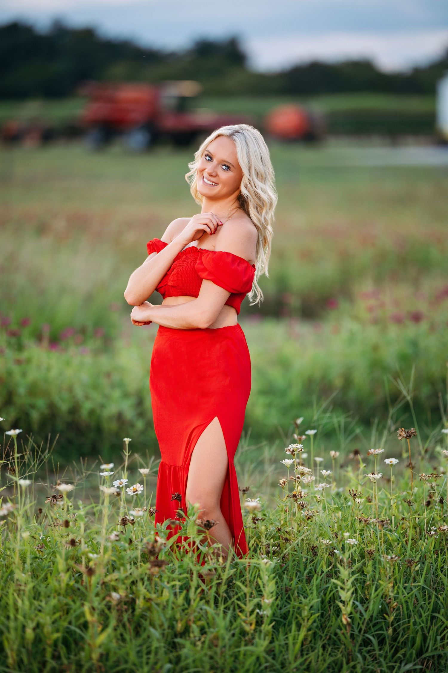 A person in a vibrant red dress poses gracefully in a summer meadow filled with wildflowers and tall grass.