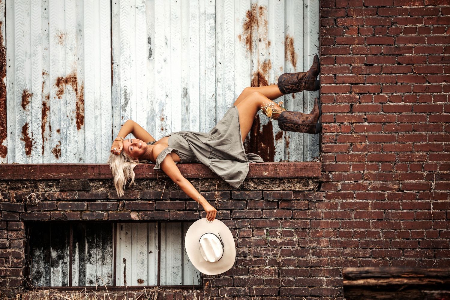 A dancer in a flowing dress performs a dramatic pose against a rustic brick wall and weathered door while holding a hat.