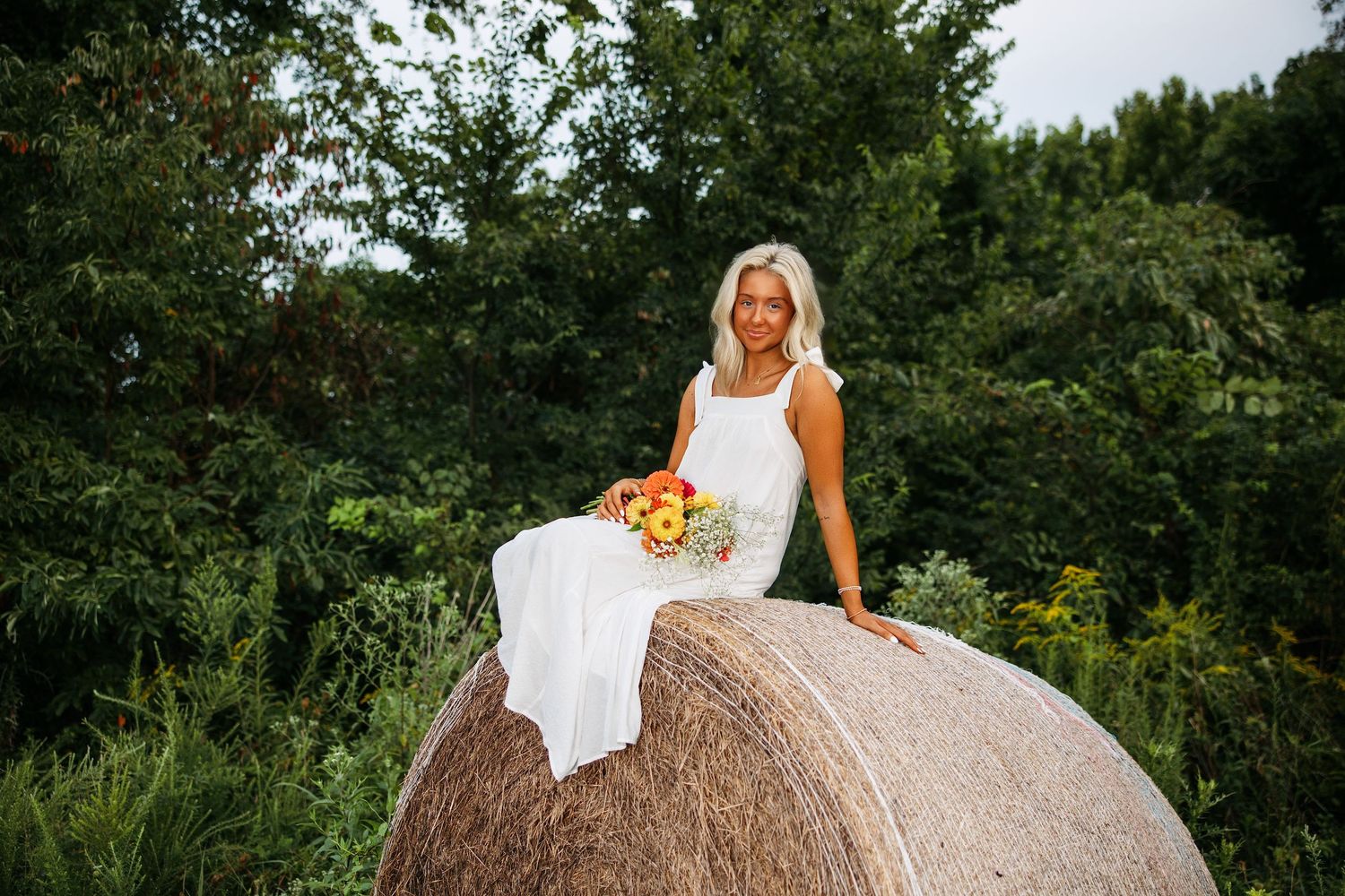 A person in a white dress poses elegantly on top of a large round hay bale in a green summer field.