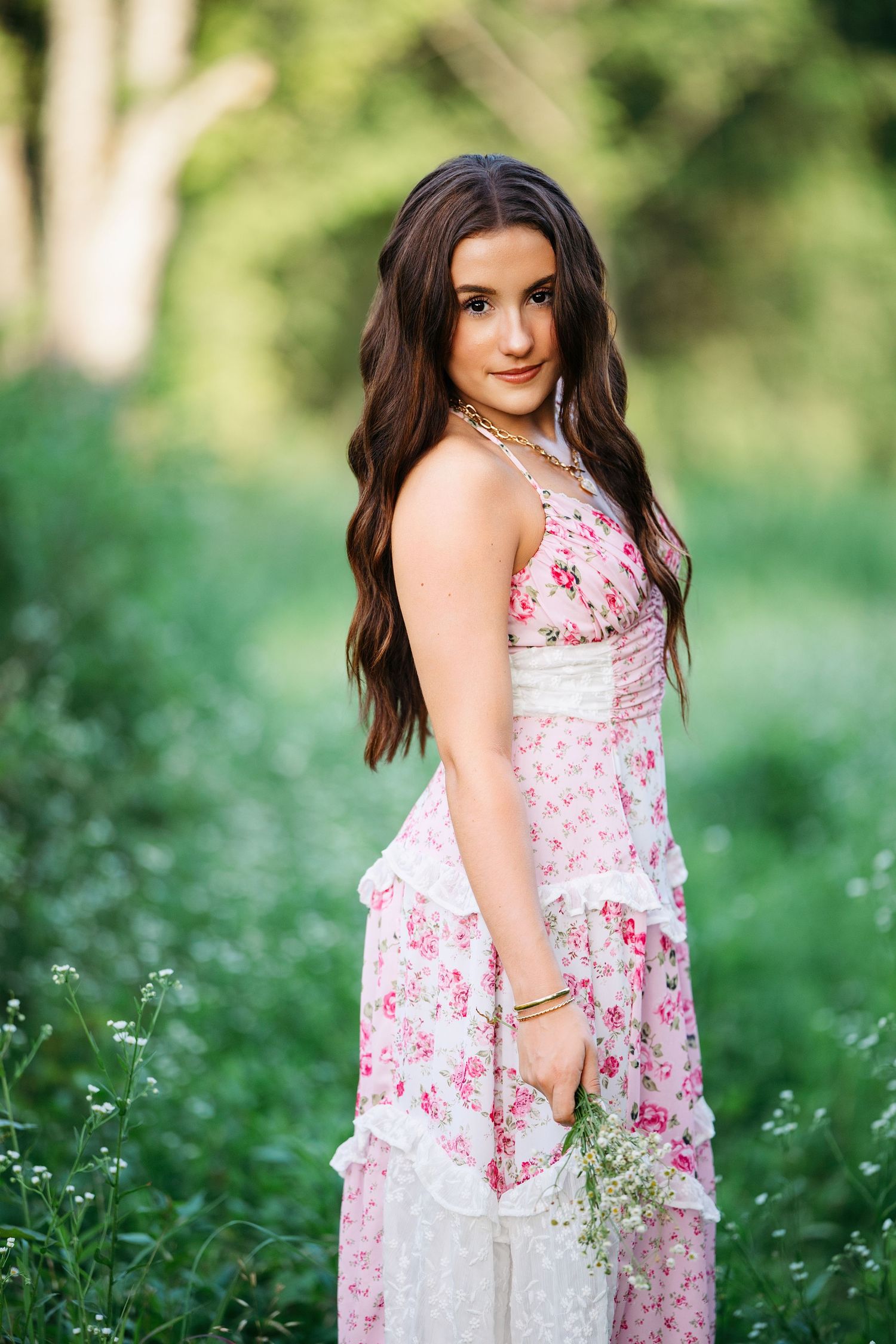 A person in a white and pink floral dress poses outdoors in a field of wildflowers during a summer photoshoot.