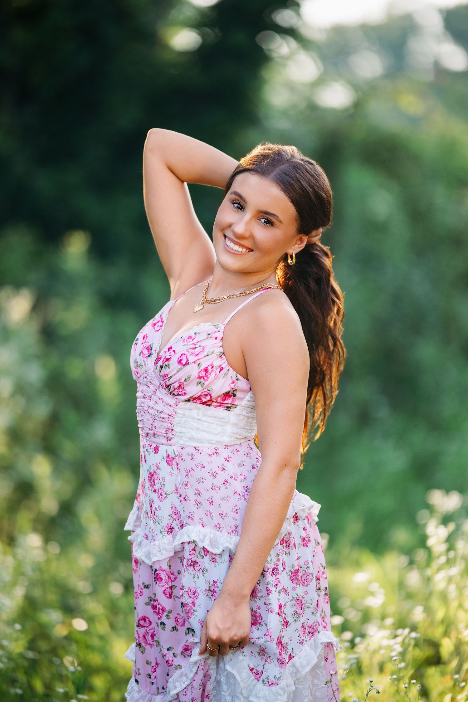 A person in a white and pink floral dress poses playfully in a sunny outdoor setting with greenery in the background.