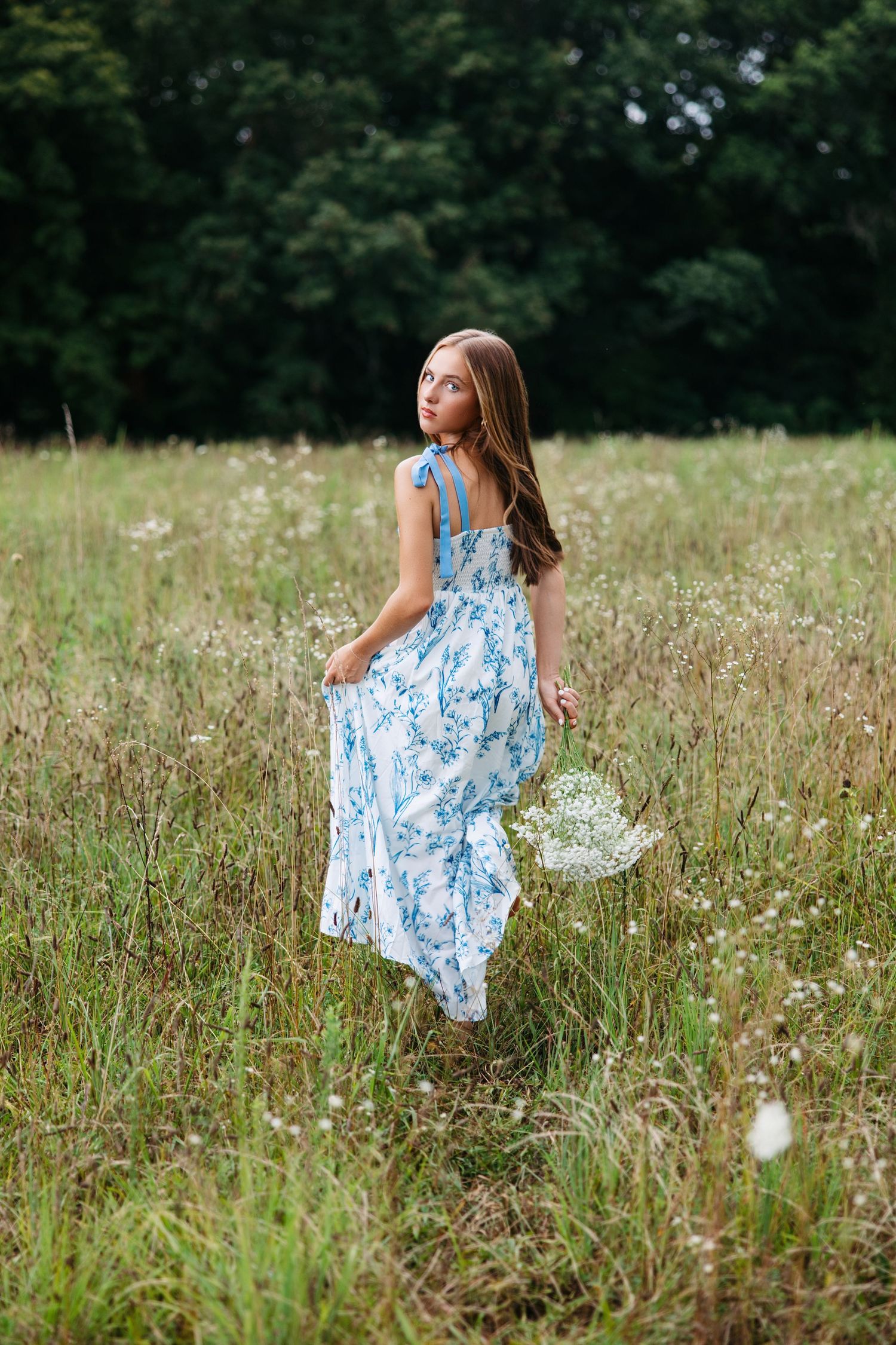 A woman in a flowing blue floral dress walks through a summer meadow holding wildflowers at sunset.