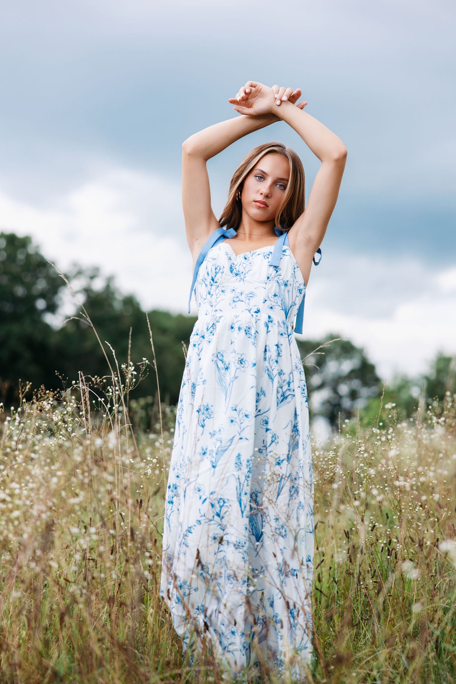 A figure in a flowing white and blue floral dress poses gracefully in a summer meadow with tall grass and wildflowers.