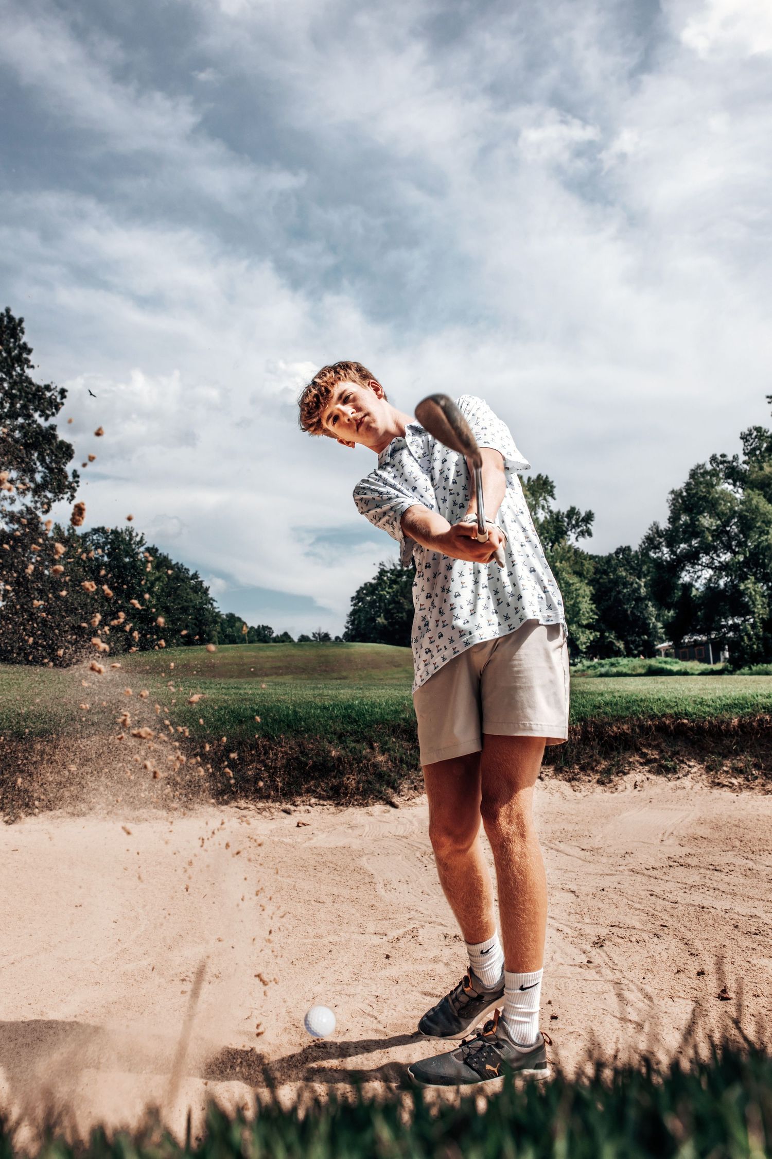 A person wearing shorts and a patterned shirt kicks up sand on a cloudy day at a golf course.