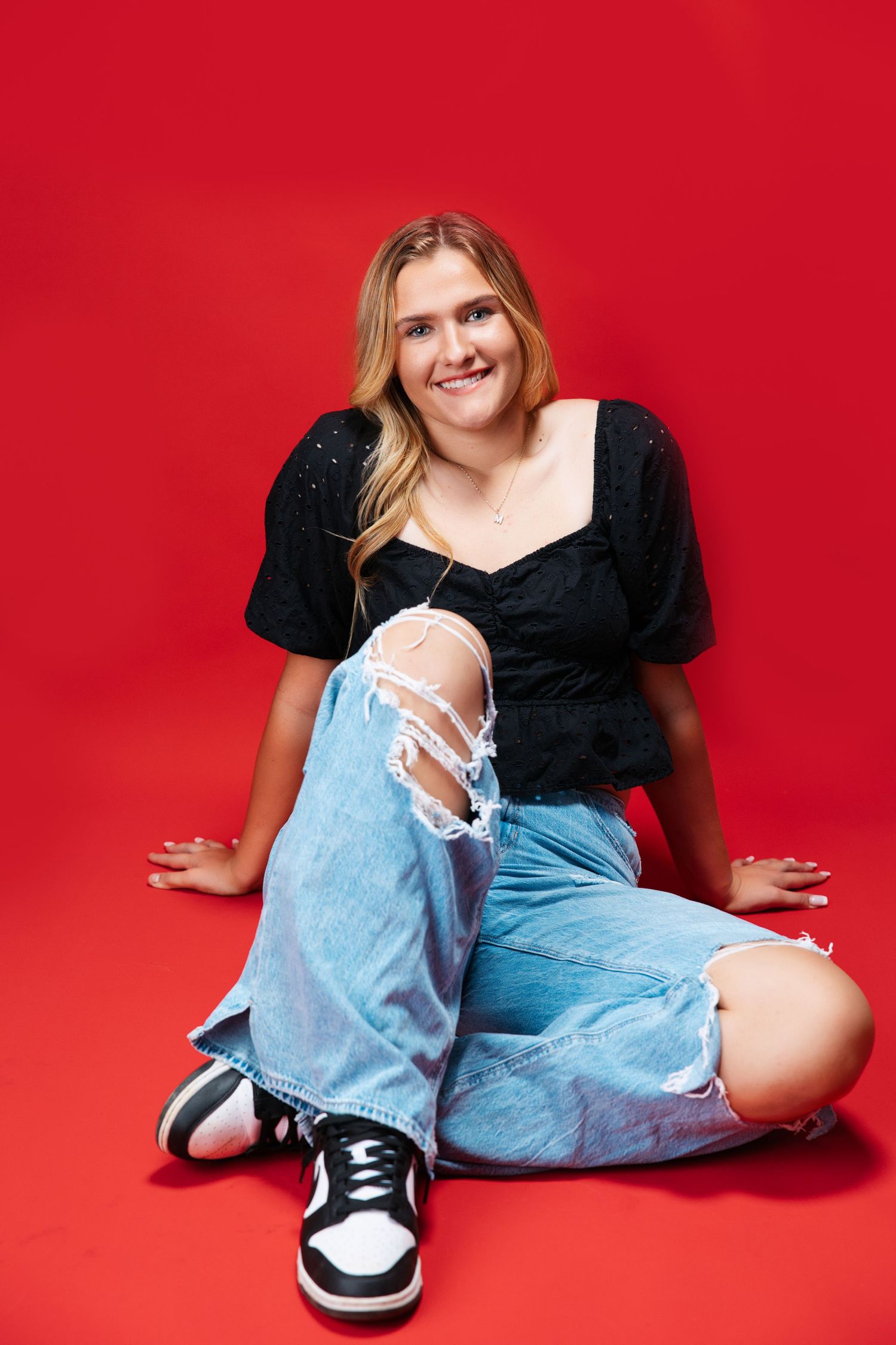 Casual pose in black top and distressed jeans against vibrant red background. Middle TN senior photographer.