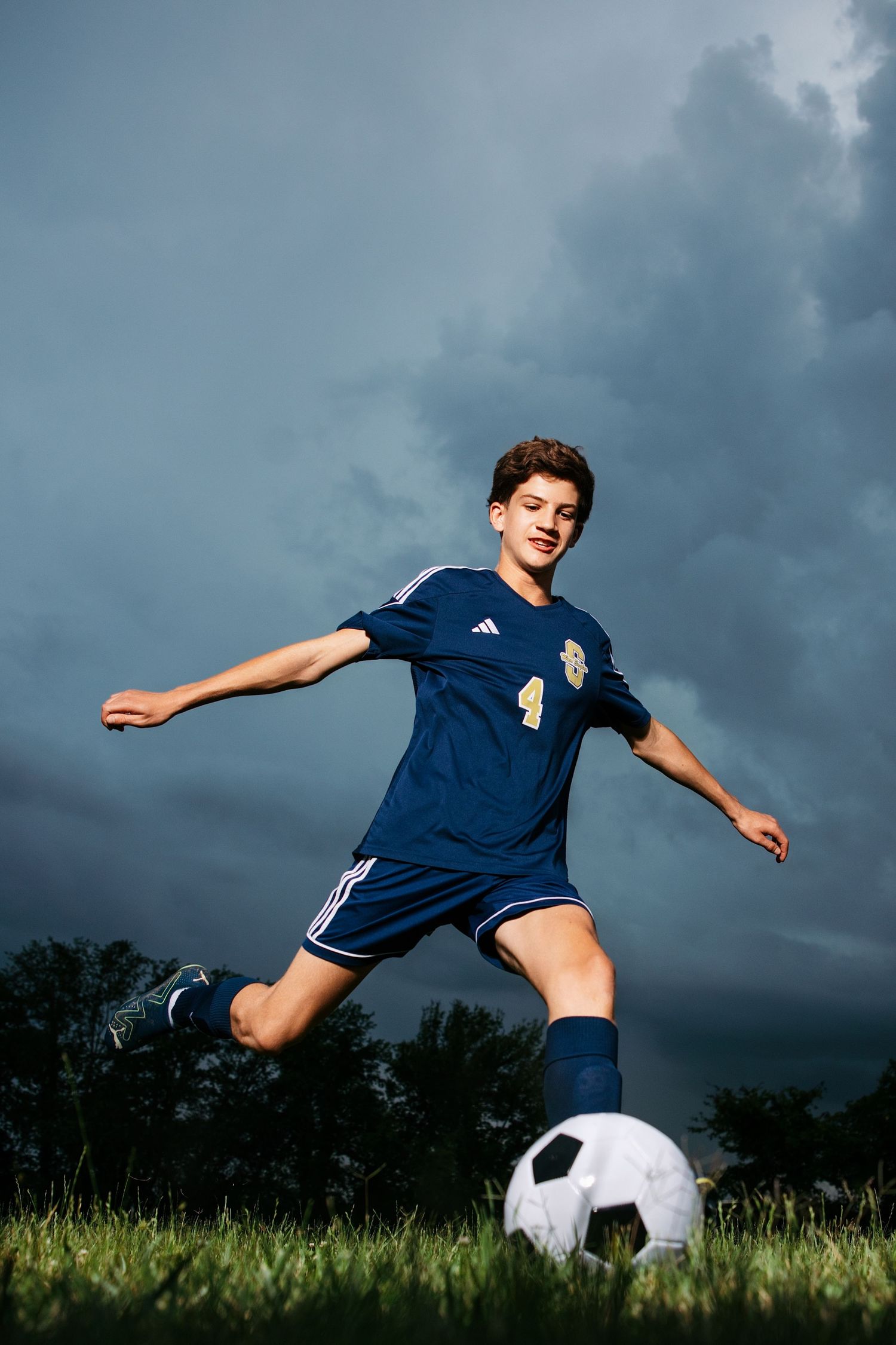A young soccer player in a navy blue jersey kicks a white ball against a dramatic stormy sky background.