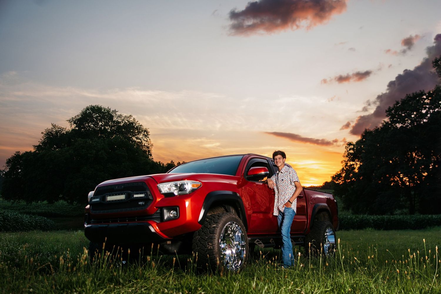 A red sports car parked in a grassy field during golden hour sunset with dramatic clouds in the sky.
