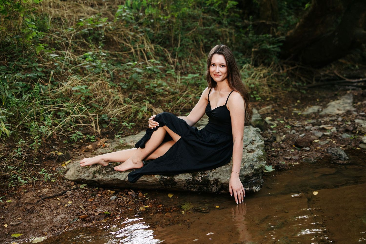 A person in a black dress poses elegantly near a creek in a forest setting during a photo session.