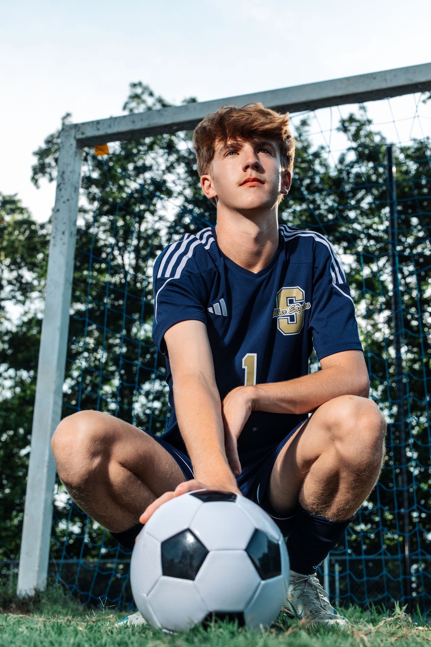 Soccer player in blue uniform crouches with ball by goal post during outdoor training session.