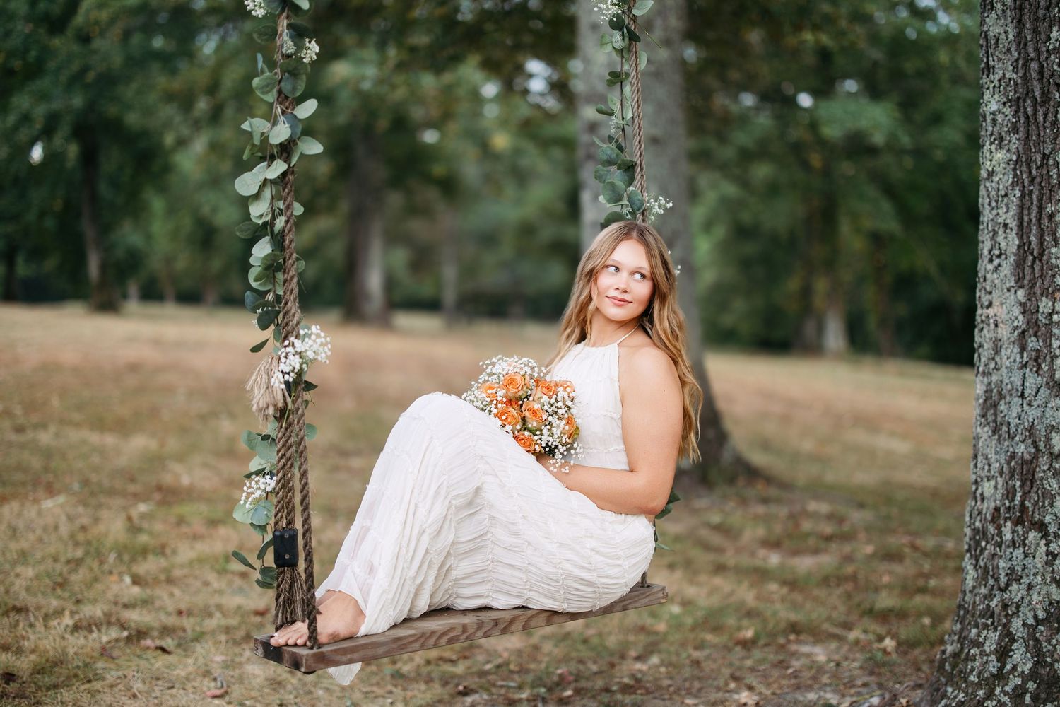 A person in a white dress sits on a rustic wooden swing adorned with floral vines in an outdoor setting.