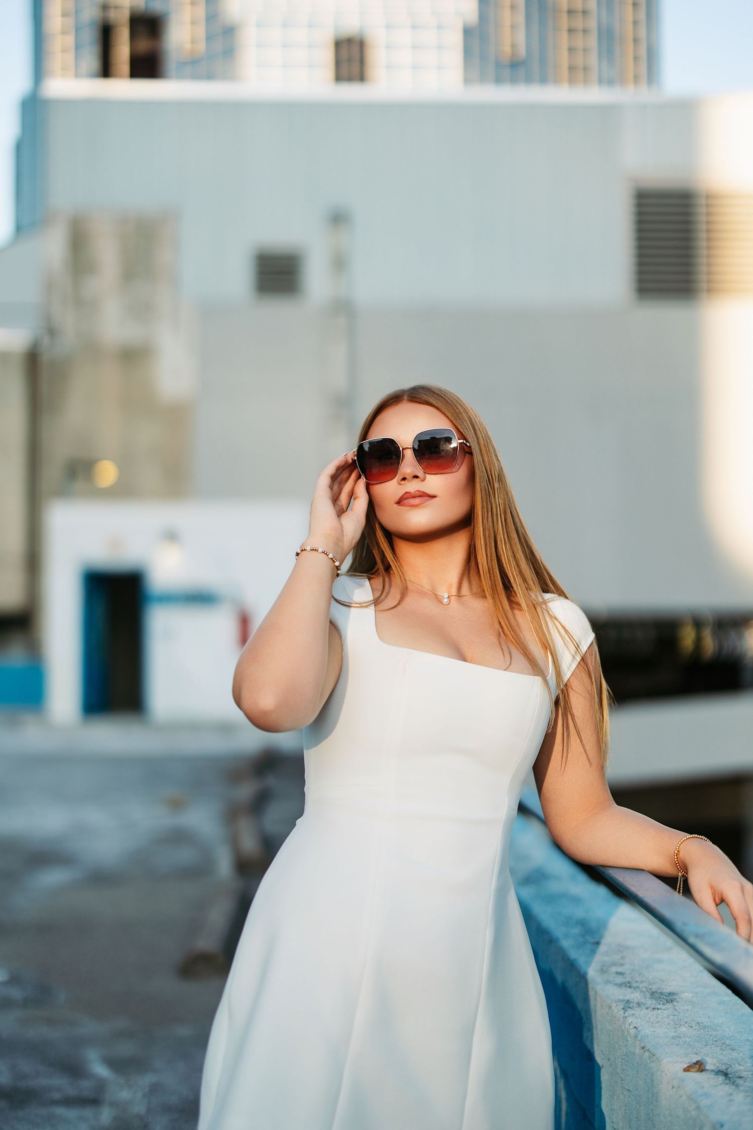 A person in a white summer dress and sunglasses poses outdoors against an urban backdrop during golden hour.