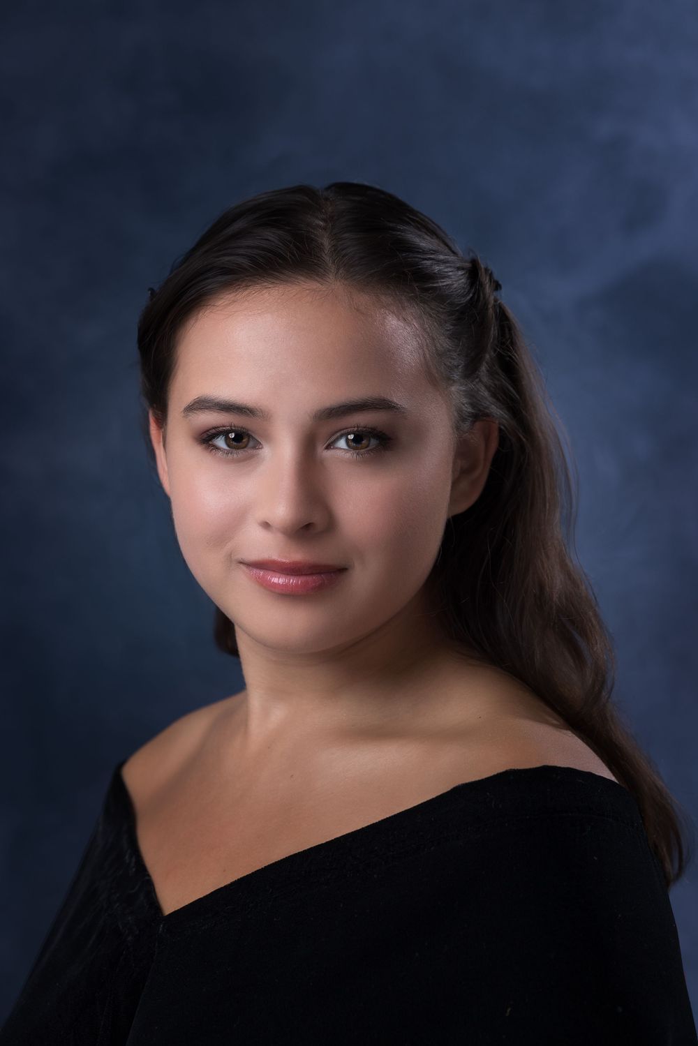 Young woman with dark hair in off-shoulder black top poses for a professional portrait against a blue backdrop.