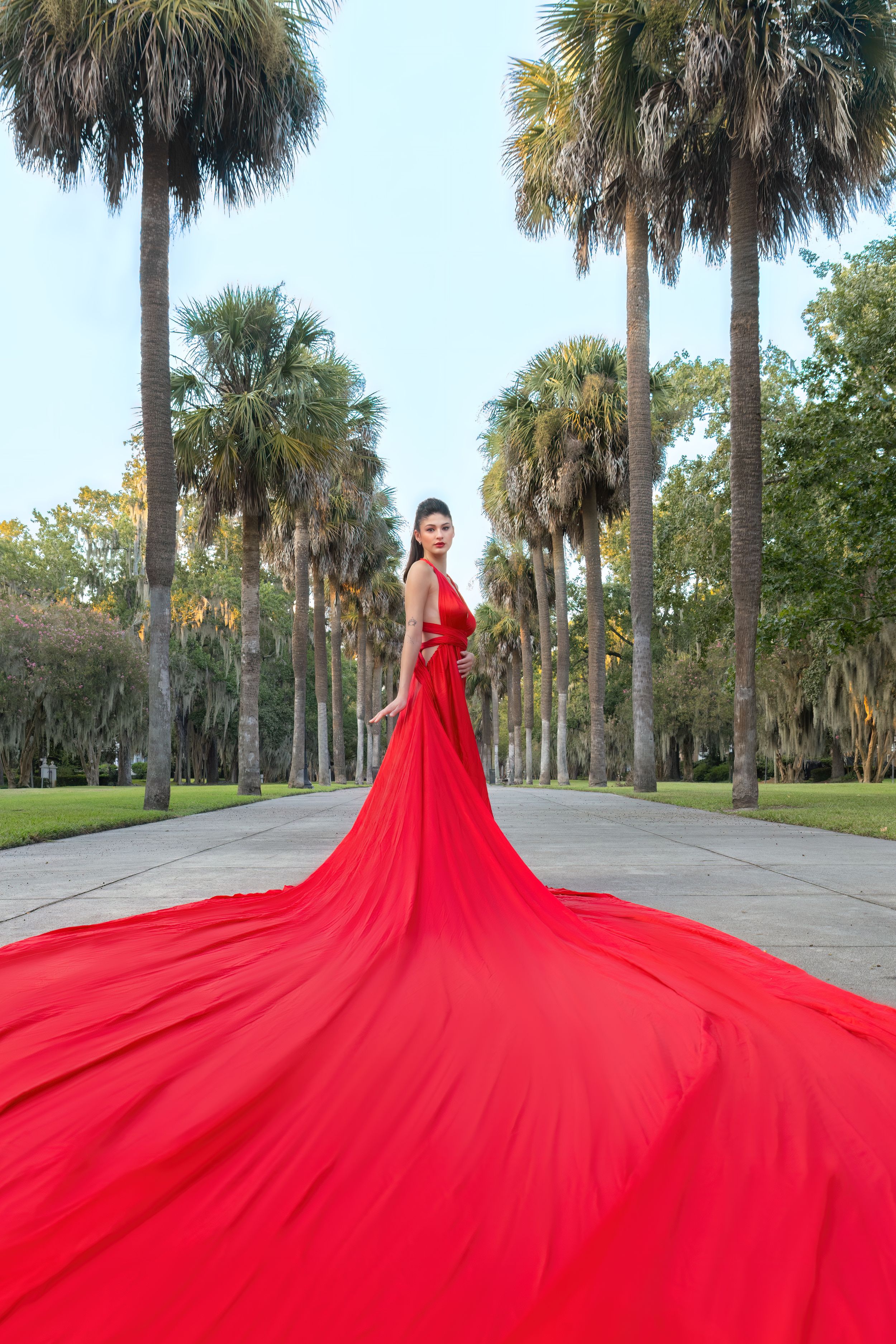 Woman in flowing red gown with long train poses among tall palm trees on a scenic pathway.