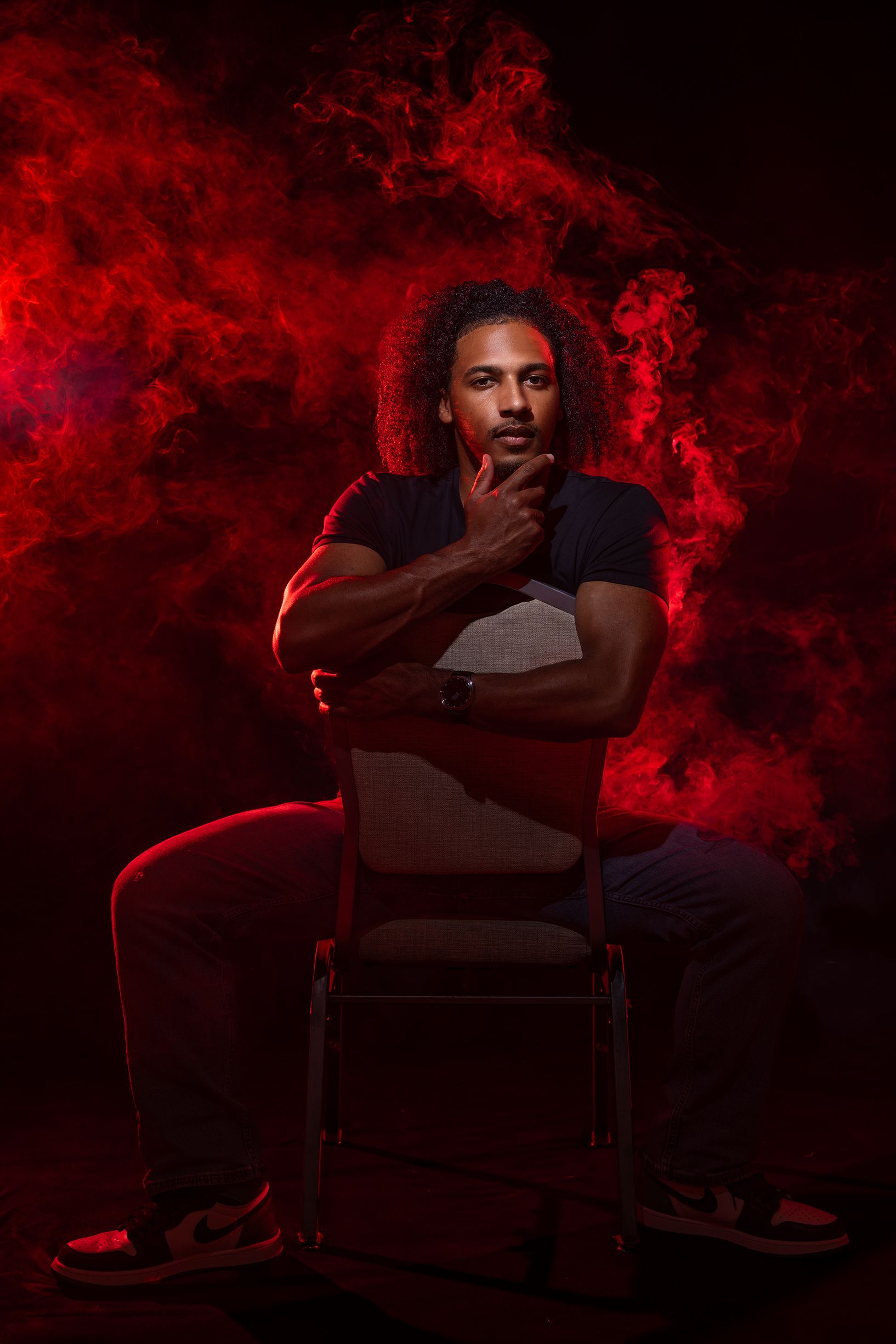 Man with curly hair sits in chair, hand on chin, against dramatic red smoky dark background.