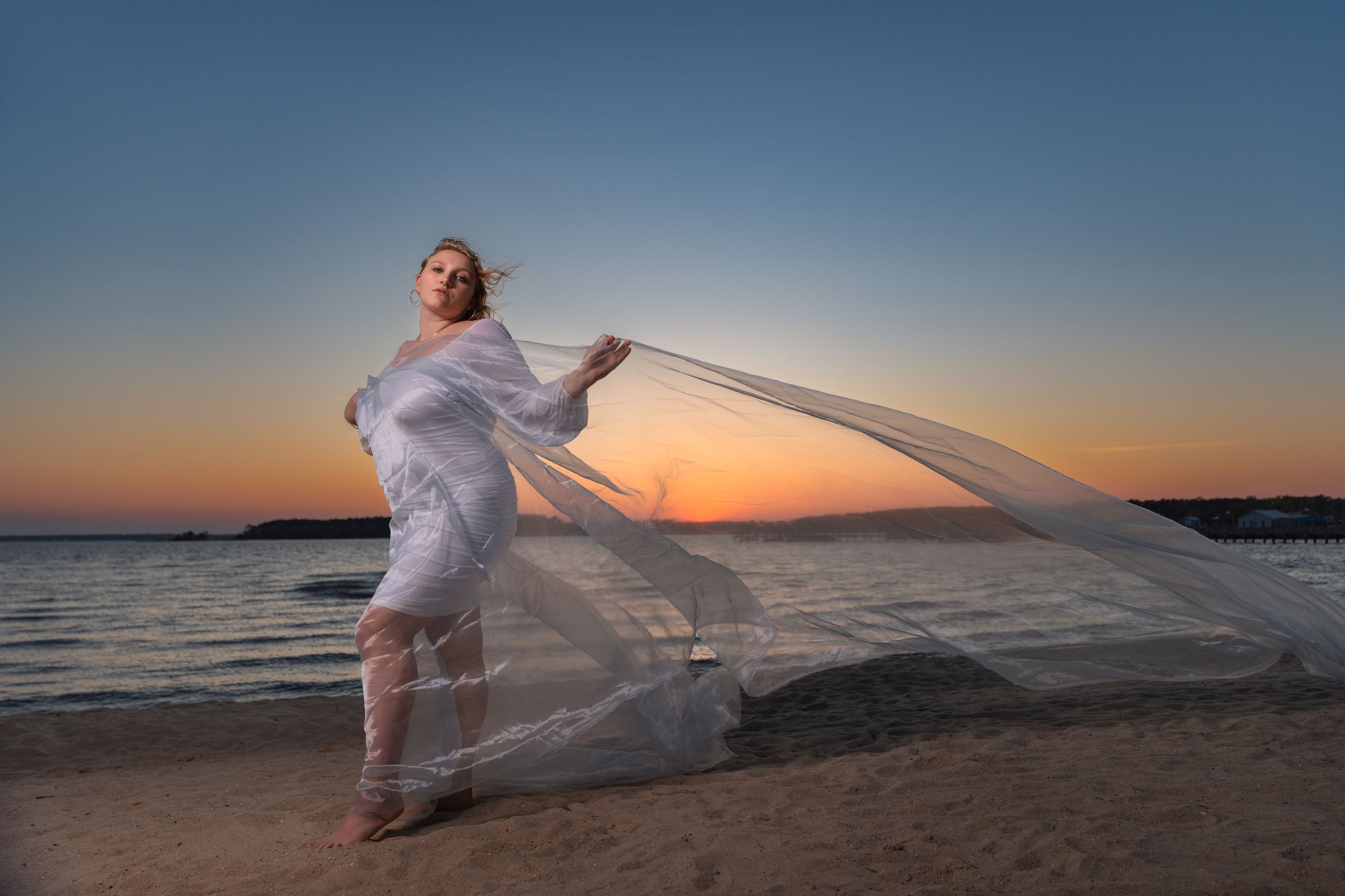 Woman in flowing white sheer fabric poses on beach at sunset with ocean backdrop.