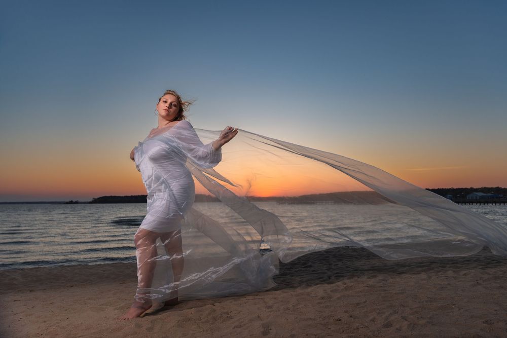 Woman in flowing white sheer fabric poses on beach at sunset with ocean backdrop.