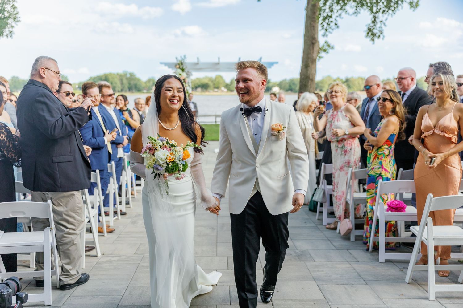 Joyful newlyweds walk down the aisle together amid smiling guests at an outdoor waterfront ceremony.