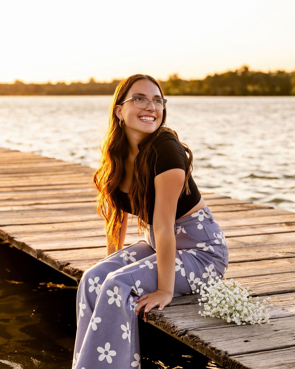 A senior girl in floral pants on a wooden dock at sunset with baby's breath flowers beside her in Omaha, NE. - by BrightSide Photography